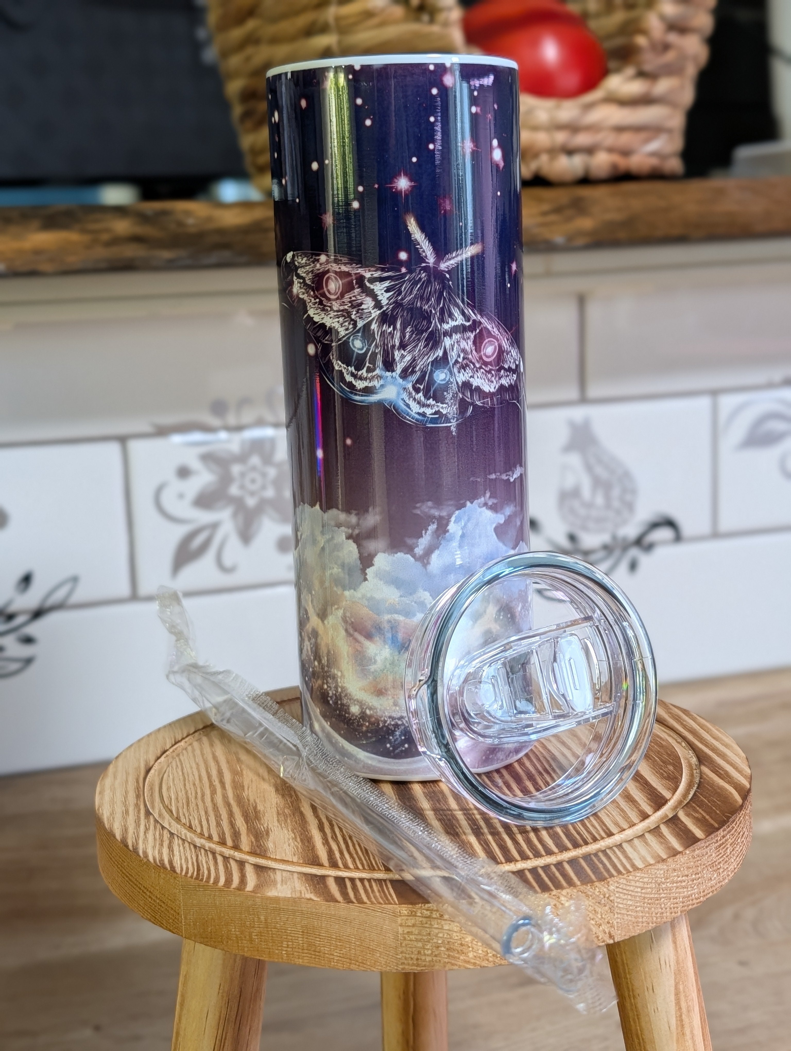 Celestial moth night sky tumbler with lid and straw beside it on wooden stool