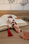 Terracotta Botanical Bookmark with red tassel tucked into open hardcover book on oak wood surface with dried terracotta leaves and eucalyptus sprig nearby