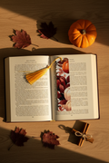 Pumpkin Harvest Bookmark with gold tassel tucked into open hardcover book on oak wood with small orange pumpkin dried burgundy leaves and cinnamon sticks