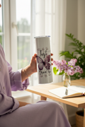 Woman in lavender linen top holding Just Breathe butterfly tumbler near bright window with lilacs and open journal