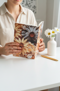 Woman holding 3D floral daisy faux leather journal showing sunflower and daisy cover at bright white desk