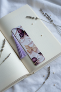 Purple Butterfly Bookmark with lavender tassel tucked into open hardcover book on white linen with scattered dried lavender sprigs and tiny pearl beads nearby
