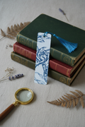 Blue Rose Etching Bookmark with teal tassel standing upright against stack of vintage hardcover books with magnifying glass and dried botanical sprigs on linen