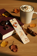 Cozy Autumn Bookmark with bronze gold tassel tucked into open hardcover book on oak wood with real mug of hot cocoa chocolate bar cinnamon sticks and dried autumn leaves